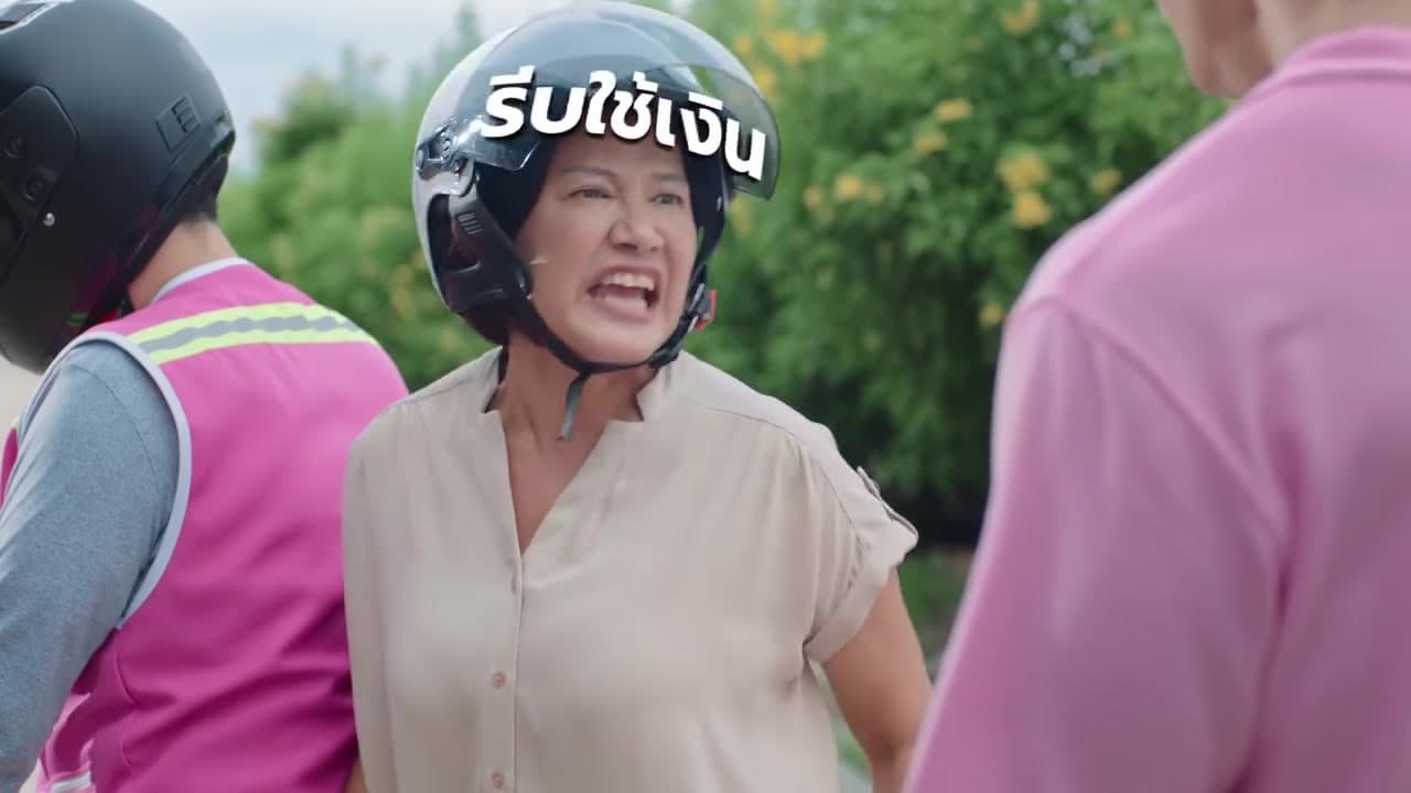 A woman in a helmet on the back of a motorbike taxi, shouting, the words “รีบใช้เงิน” (need cash fast) across her helmet
