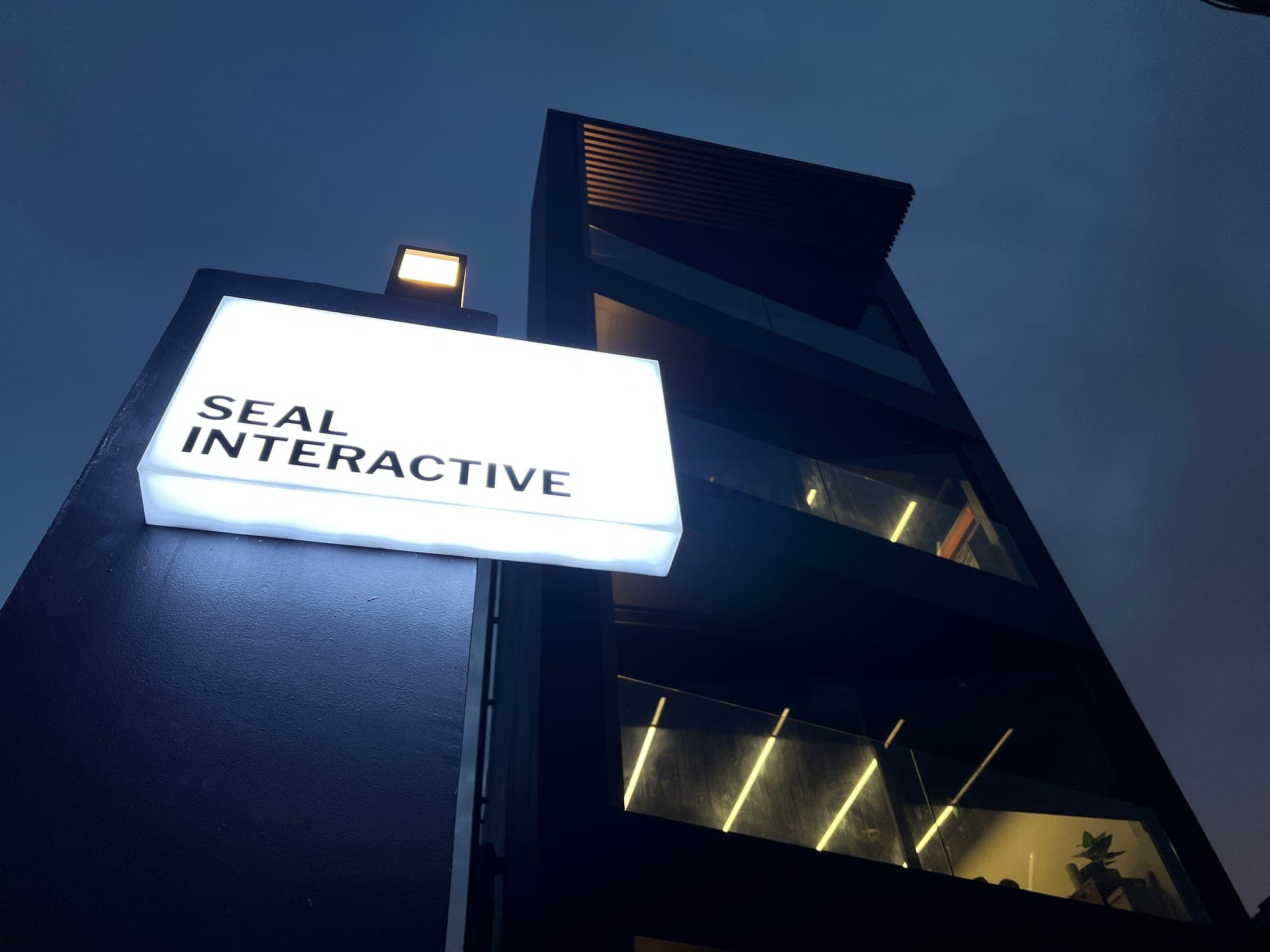 ออฟฟิศ Seal Interactive ยามพลบค่ำ — อาคารสีดำกับป้ายไฟกล่องสีขาว